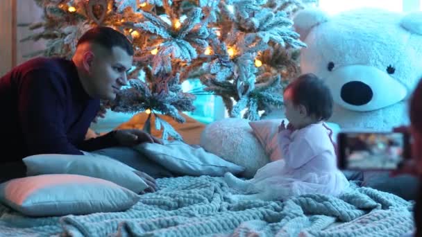 Feliz familia sonriente cerca del árbol de Navidad. mujer toma fotos en el teléfono de un niño — Vídeos de Stock