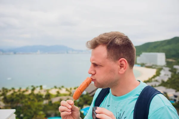 Man looking sausage with curious thinking face. Having breakfast eating holding hot dog.