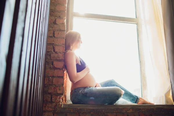 Joven hermosa mujer embarazada sentada en el alféizar de la ventana en casa —  Fotos de Stock