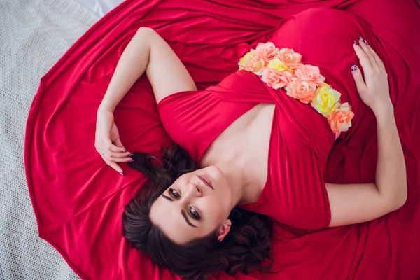 Güzel bir hamile kadın siyah iç çamaşırı üzerinde gri bir peçe yatıyor. Yatağın taze çiçeklerle bezenmiştir. Kız sarışın, o gülümsüyor vardır. Yukarıdan görüntülemek.