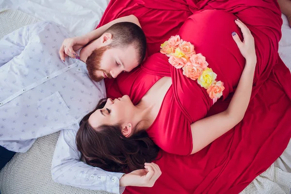 Uma bela mulher grávida de roupa interior preta está em um véu cinza. A cama está decorada com flores frescas. A menina tem cabelo loiro, sorri. Vista de cima . — Fotografia de Stock