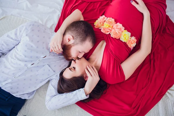 Eine schöne schwangere Frau in schwarzer Unterwäsche liegt auf einem grauen Schleier. Das Bett ist mit frischen Blumen dekoriert. Das Mädchen hat blonde Haare, sie lächelt. Blick von oben. — Stockfoto