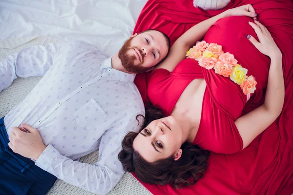 Eine schöne schwangere Frau in schwarzer Unterwäsche liegt auf einem grauen Schleier. Das Bett ist mit frischen Blumen dekoriert. Das Mädchen hat blonde Haare, sie lächelt. Blick von oben. — Stockfoto