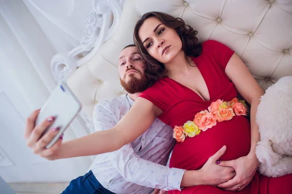 Um marido e uma esposa grávida fazem selfie sentados no sofá. Eles amam-se. Eles estão em casa . — Fotografia de Stock