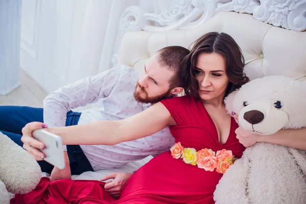 Um marido e uma esposa grávida fazem selfie sentados no sofá. Eles amam-se. Eles estão em casa . — Fotografia de Stock