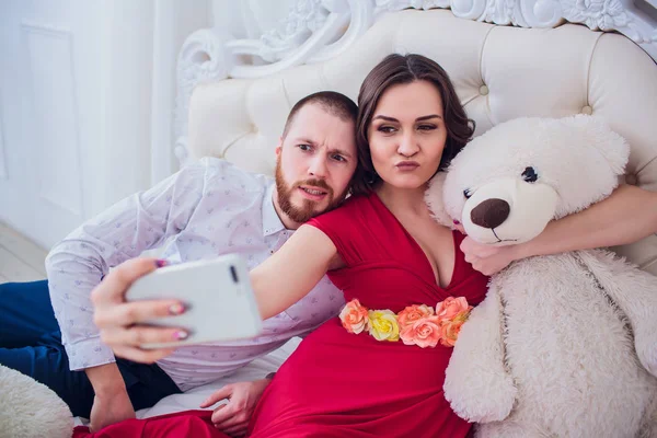 Um marido e uma esposa grávida fazem selfie sentados no sofá. Eles amam-se. Eles estão em casa . — Fotografia de Stock