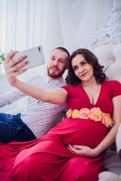 Manžel a těhotnou manželku udělat selfie sedí na gauči. Milují jeden druhého. Oni jsou doma. — Stock fotografie