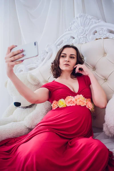 Feliz sorrindo mulher grávida sentada na grama e faz auto-retrato no smartphone. a mulher grávida faz selfie . — Fotografia de Stock