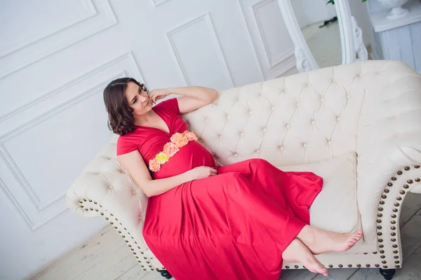 Těhotná žena v červených šatech ležela na pohovce — Stock fotografie