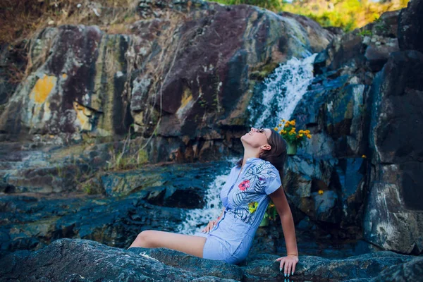 Jovem com cabelo preto nas pedras contra o fundo de uma cachoeira correndo — Fotografia de Stock
