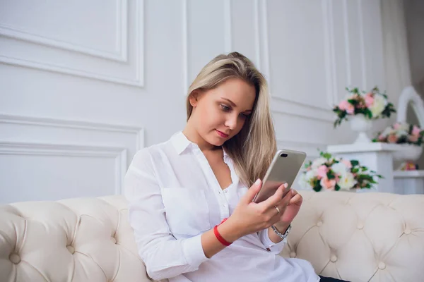 Retrato de close-up de uma mulher loira sentada no sofá e usando seu telefone inteligente. Mulher bonita tocando a tela e fazendo chamada . — Fotografia de Stock
