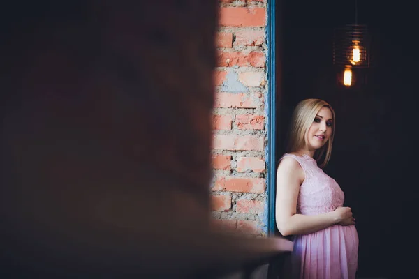Casualmente vestido mulher grávida atraente em pé em casa, olhando câmera e sorrindo enquanto — Fotografia de Stock