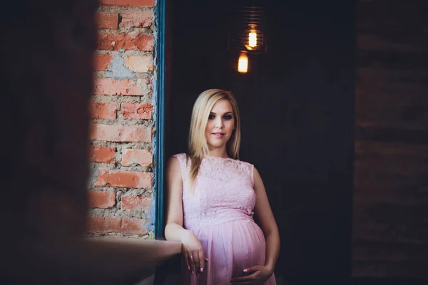 Lässig gekleidete attraktive schwangere Frau, die zu Hause steht, Kamera schaut und lächelt, während — Stockfoto