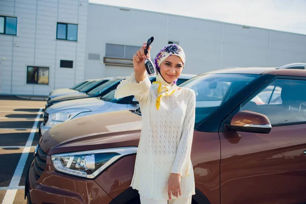 Propiedad y el concepto de la gente - mujer musulmana en hijab con la llave del coche sobre el fondo del espectáculo de coches. mujer feliz tomando la llave del coche del distribuidor en el espectáculo auto o salón —  Fotos de Stock