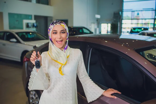 property and people concept - muslim woman in hijab with car key over car show background. happy woman taking car key from dealer in auto show or salon fools around