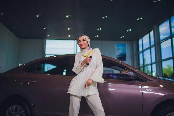 Propiedad y el concepto de la gente - mujer musulmana en hijab con la llave del coche sobre el fondo del espectáculo de coches. mujer feliz tomando la llave del coche del distribuidor en el espectáculo auto o salón —  Fotos de Stock