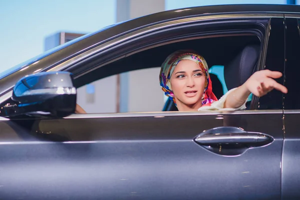 property and people concept - muslim woman in hijab with car key over car show background. happy woman taking car key from dealer in auto show or salon
