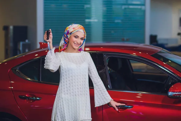 Propiedad y el concepto de la gente - mujer musulmana en hijab con la llave del coche sobre el fondo del espectáculo de coches. mujer feliz tomando la llave del coche del distribuidor en el espectáculo auto o salón — Foto de Stock