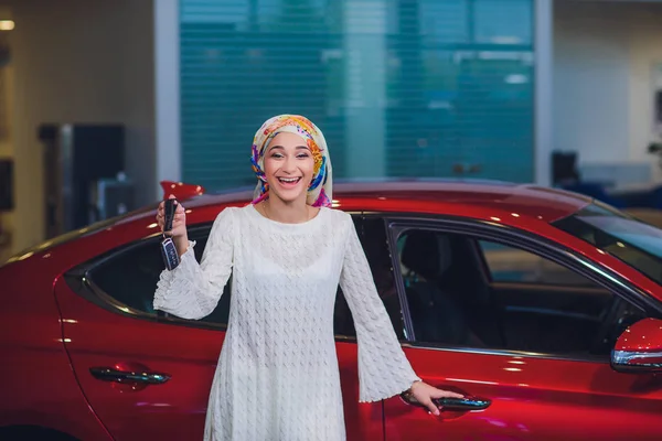 property and people concept - muslim woman in hijab with car key over car show background. happy woman taking car key from dealer in auto show or salon