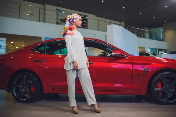 property and people concept - muslim woman in hijab with car key over car show background. happy woman taking car key from dealer in auto show or salon.