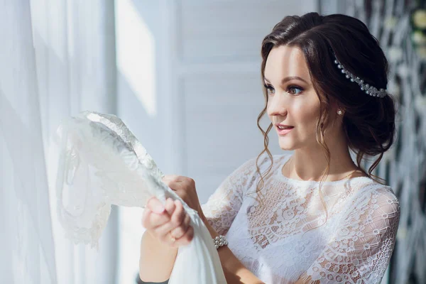 Bella sposa bruna in vestaglia di seta e velo di pizzo che tiene l'abito da sposa su un appendiabiti vicino alla finestra . — Foto Stock