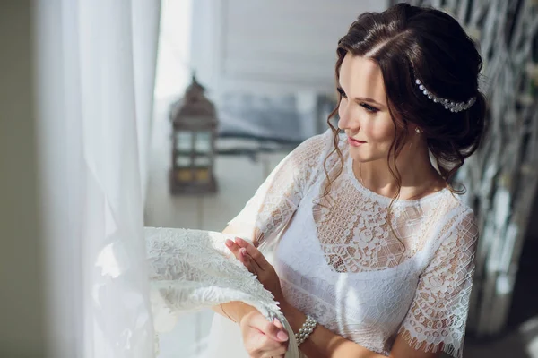 Bella sposa bruna in vestaglia di seta e velo di pizzo che tiene l'abito da sposa su un appendiabiti vicino alla finestra . — Foto Stock