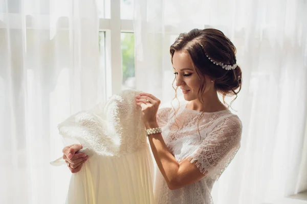 Mooie brunette bruid in zijde badjas en lacy sluier trouwjurk te houden op een hanger in de buurt van het venster. — Stockfoto