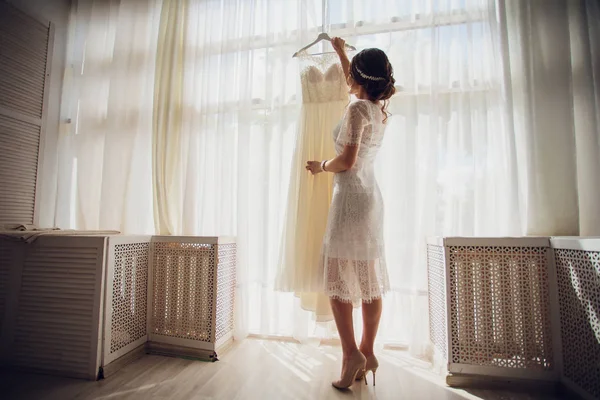 Hübsche brünette Braut in seidenem Morgenmantel und Spitzenschleier mit Brautkleid auf einem Kleiderbügel am Fenster. — Stockfoto