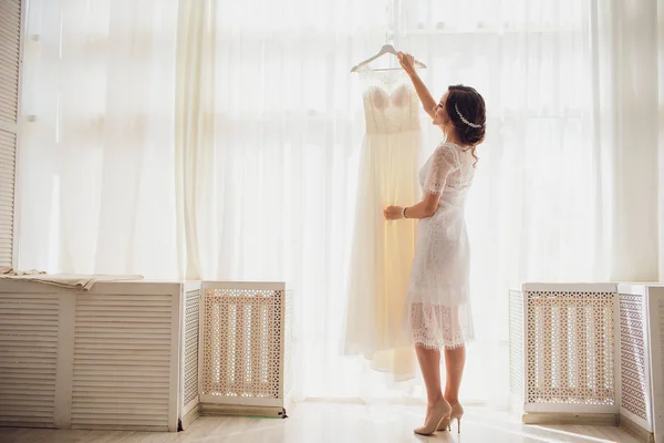 Hübsche brünette Braut in seidenem Morgenmantel und Spitzenschleier mit Brautkleid auf einem Kleiderbügel am Fenster. — Stockfoto