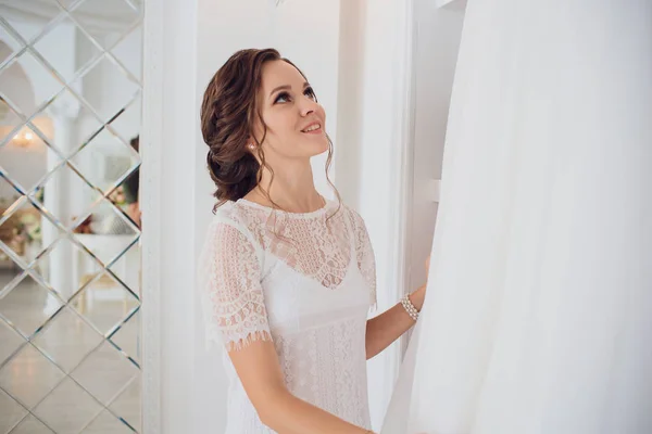 Гарненька брюнетка наречена в шовковій сукні і мереживна вуаль тримає весільну сукню на вішалці біля вікна . — стокове фото