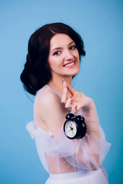 Hermosa chica encantadora en vestido de novia blanco, siendo tarde en la mañana, y sosteniendo en su mano reloj despertador retro y anillo — Foto de Stock