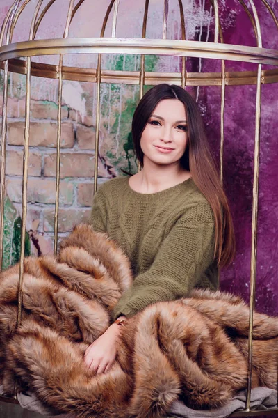 Beautiful girl in green sweater is sitting on big cozy carpet and holding boxes of gifts for her beloved. On the back there are a white fireplace covered sits in a cage — Stock Photo, Image