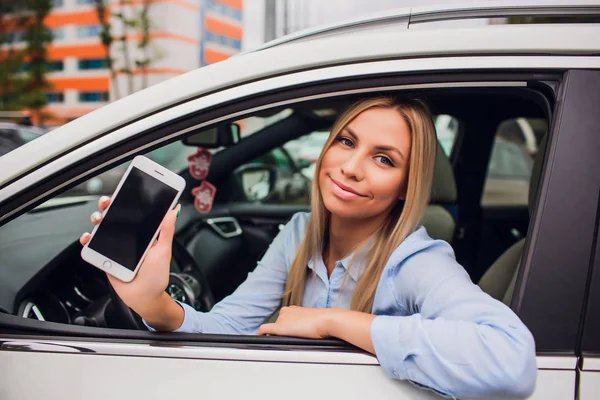Mujer rubia feliz mostrando teléfono inteligente por la ventana del coche. Centrarse en el teléfono móvil . —  Fotos de Stock