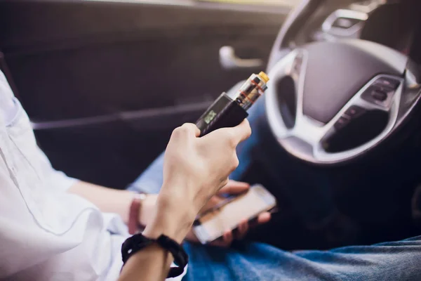 Vista desde el lado de un joven fumando un cigarrillo electrónico mientras conduce su coche en una calle urbana Sostiene y ajusta el poder del cigarrillo electrónico — Foto de Stock