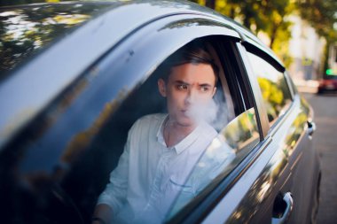 View from side young man smoking an e-cigarette as he drives his car on an urban street. car driver peeps out of the car and smokes clipart