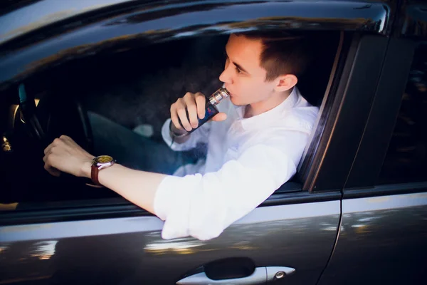 Vista desde un lado joven fumando un cigarrillo electrónico mientras conduce su coche en una calle urbana. conductor del coche se asoma fuera del coche y fuma — Foto de Stock