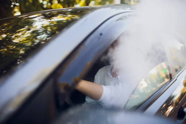 Visa från sida ung man röka en e-cigarett som han kör sin bil på en urban street. bilförare peeps ur bil och röker — Stockfoto