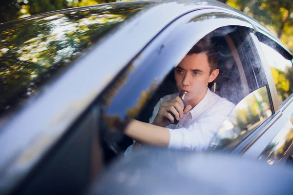 Vista desde un lado joven fumando un cigarrillo electrónico mientras conduce su coche en una calle urbana. conductor del coche se asoma fuera del coche y fuma — Foto de Stock
