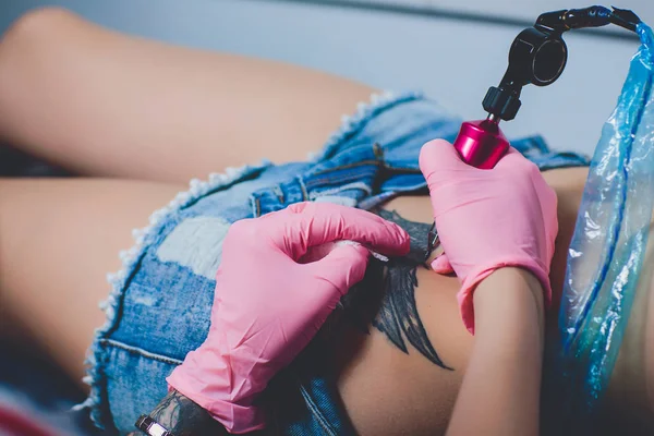 Tatuering artist, tatuering på hennes mage, rosa skrivmaskin, specialist i rosa handskar, vit bakgrund. — Stockfoto