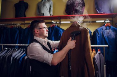 terzi keser ve bir takım elbise diker. Terzilik dükkanı genç bir erkekte erkek giyim yapar. Ismarlama giyim Atölyesi. onun elinde Elektrikli Ütü tutar. ıslak-ısıl işlem