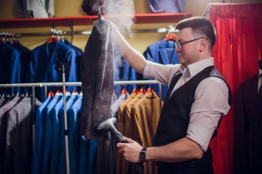 terzi keser ve bir takım elbise diker. Terzilik dükkanı genç bir erkekte erkek giyim yapar. Ismarlama giyim Atölyesi. onun elinde Elektrikli Ütü tutar. ıslak-ısıl işlem