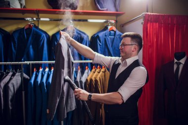 terzi keser ve bir takım elbise diker. Terzilik dükkanı genç bir erkekte erkek giyim yapar. Ismarlama giyim Atölyesi. onun elinde Elektrikli Ütü tutar. ıslak-ısıl işlem