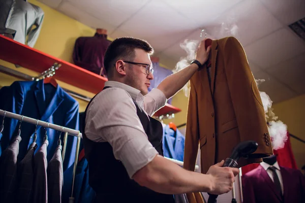 tailor cuts and sews a suit. A young man in the tailoring shop makes mens clothing. Custom-made clothing workshop. holds in her hand an electric iron. wet-heat treatment