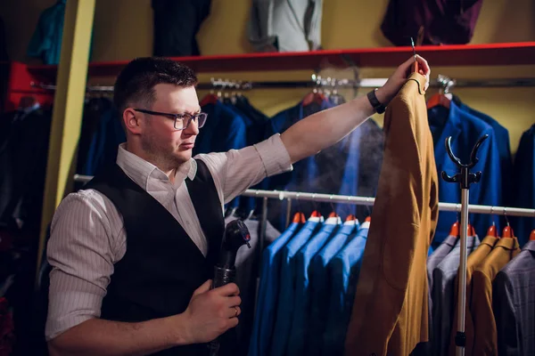 Személyre szabott darabok és sews öltönyt. Egy fiatal férfi a szabászati boltban teszi a férfi ruházat. Rendelésre készült ruházatot workshop. tart a kezében egy elektromos vasalót. Nedves hőkezelési eljárás — Stock Fotó