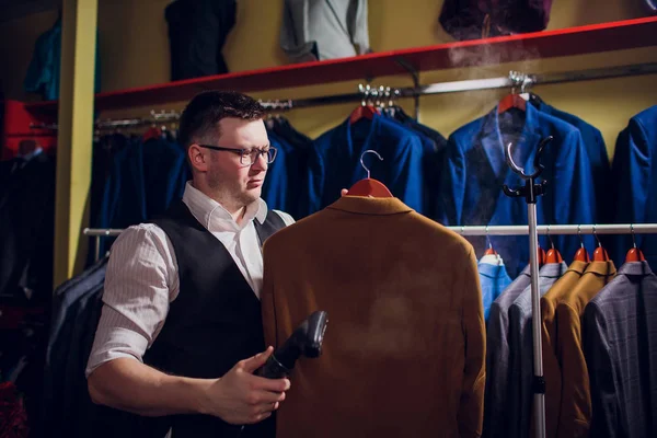 tailor cuts and sews a suit. A young man in the tailoring shop makes mens clothing. Custom-made clothing workshop. holds in her hand an electric iron. wet-heat treatment