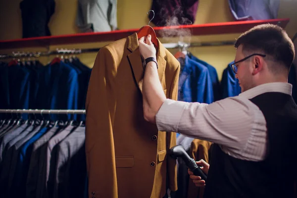 tailor cuts and sews a suit. A young man in the tailoring shop makes mens clothing. Custom-made clothing workshop. holds in her hand an electric iron. wet-heat treatment