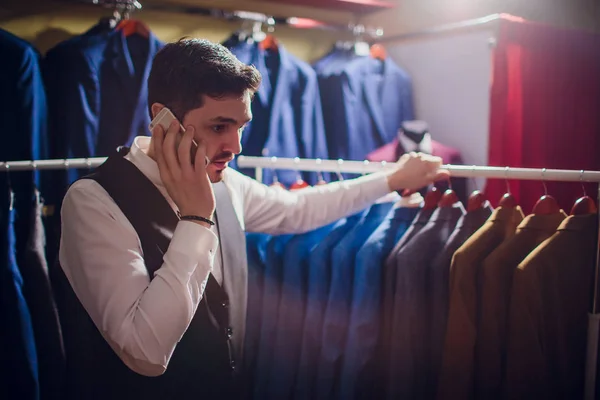 sale, shopping, fashion, communication and people concept - happy young man or businessman calling on smartphone and choosing clothes clothing store