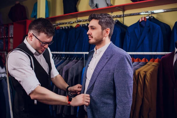 Businessman in classic vest against row suits in shop. Man helps another try on suit in clothing store