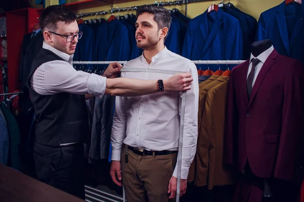 Empresário em colete clássico contra ternos de linha na loja. Homem ajuda outra tentativa em terno na loja de roupas — Fotografia de Stock