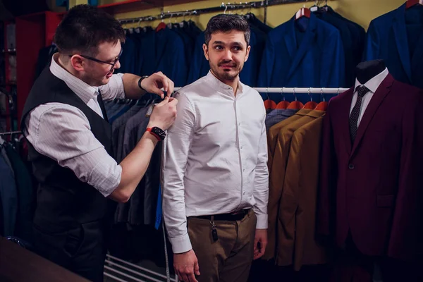 Empresario en chaleco clásico contra los trajes de fila en la tienda. El hombre ayuda a probar otro traje en la tienda de ropa — Foto de Stock